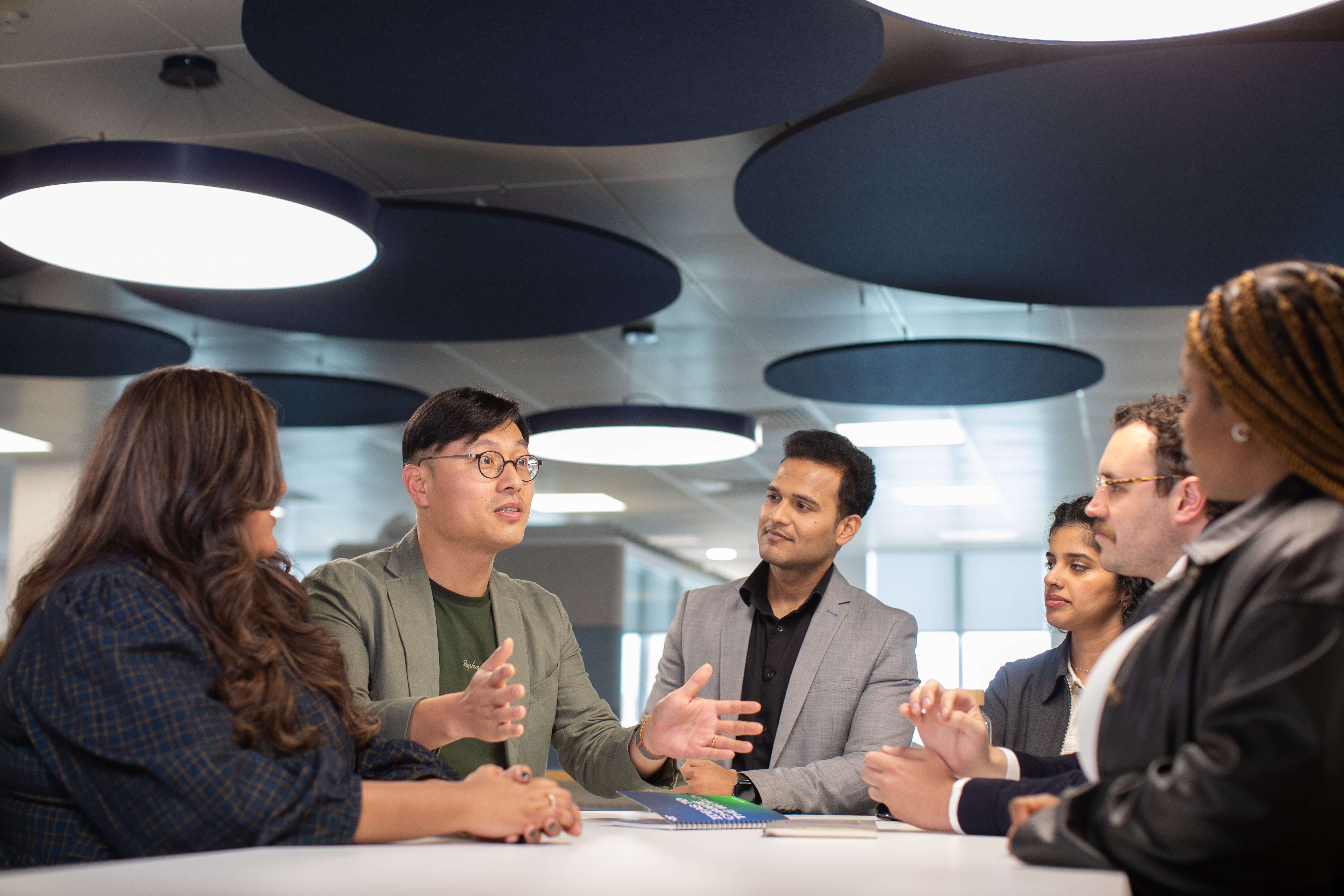 A diverse group of six professionals having a discussion around a table in a modern office setting, with circular ceiling panels above them.