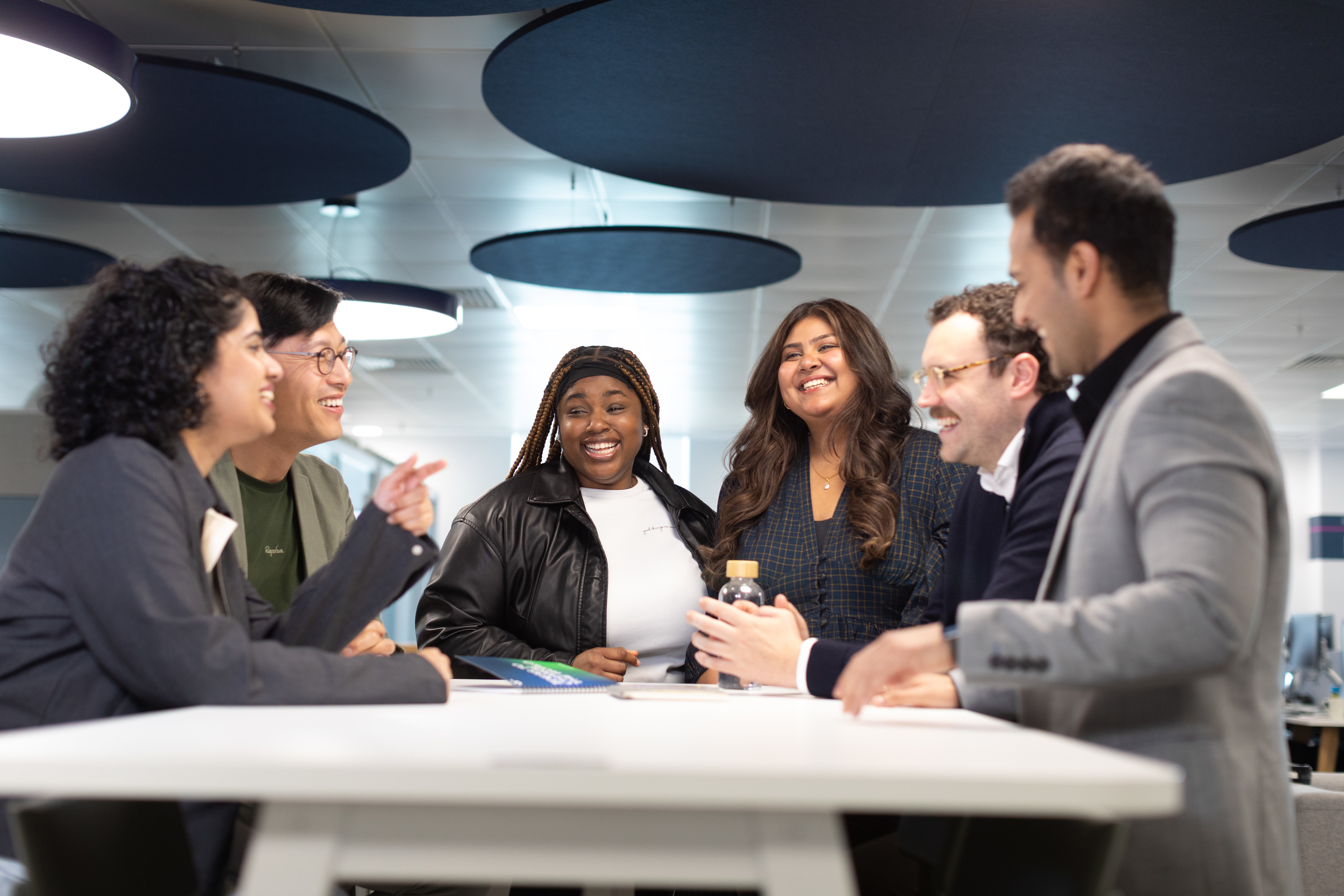 Diverse team gathered around a table in a collaborative meeting, all smiling and engaged in conversation in a contemporary workspace.