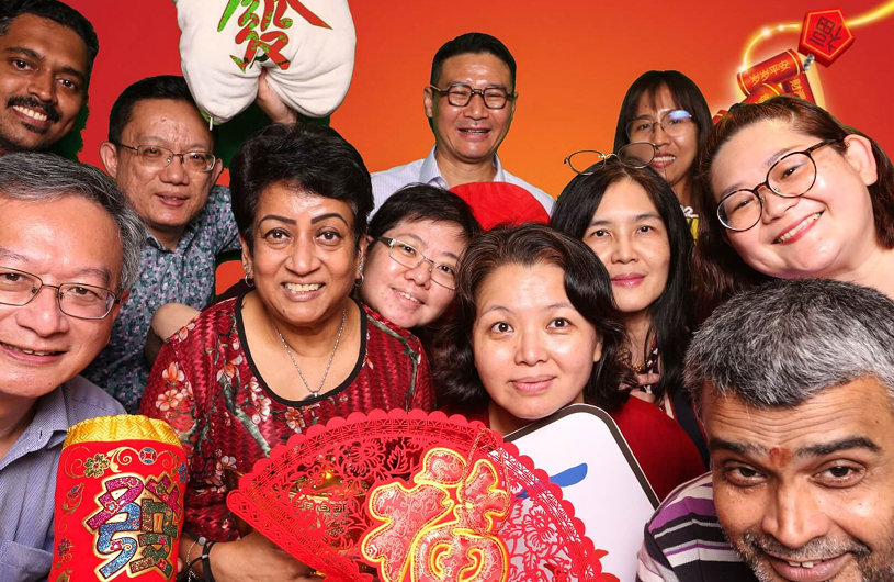 A diverse office team smiling and posing closely together in front of a festive red and orange background, celebrating Chinese New Year with traditional decorations and props.