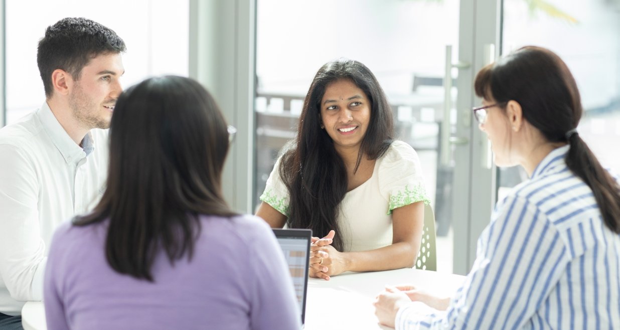 “Four people sitting around a table in a bright office, having a discussion. One woman in a white top is speaking while the others listen attentively.