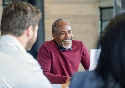 A middle-aged man with gray hair and a beard, wearing a maroon sweater, smiling and engaged in conversation with two other people whose backs are to the camera. They are indoors in a casual meeting setting.