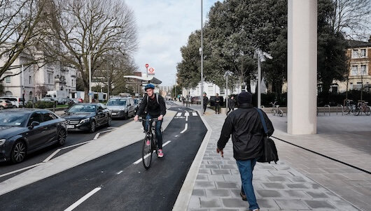 A cyclist rides on a dedicated bike lane next to a pedestrian walking on a sidewalk in an urban area with parked cars and buildings in the background.