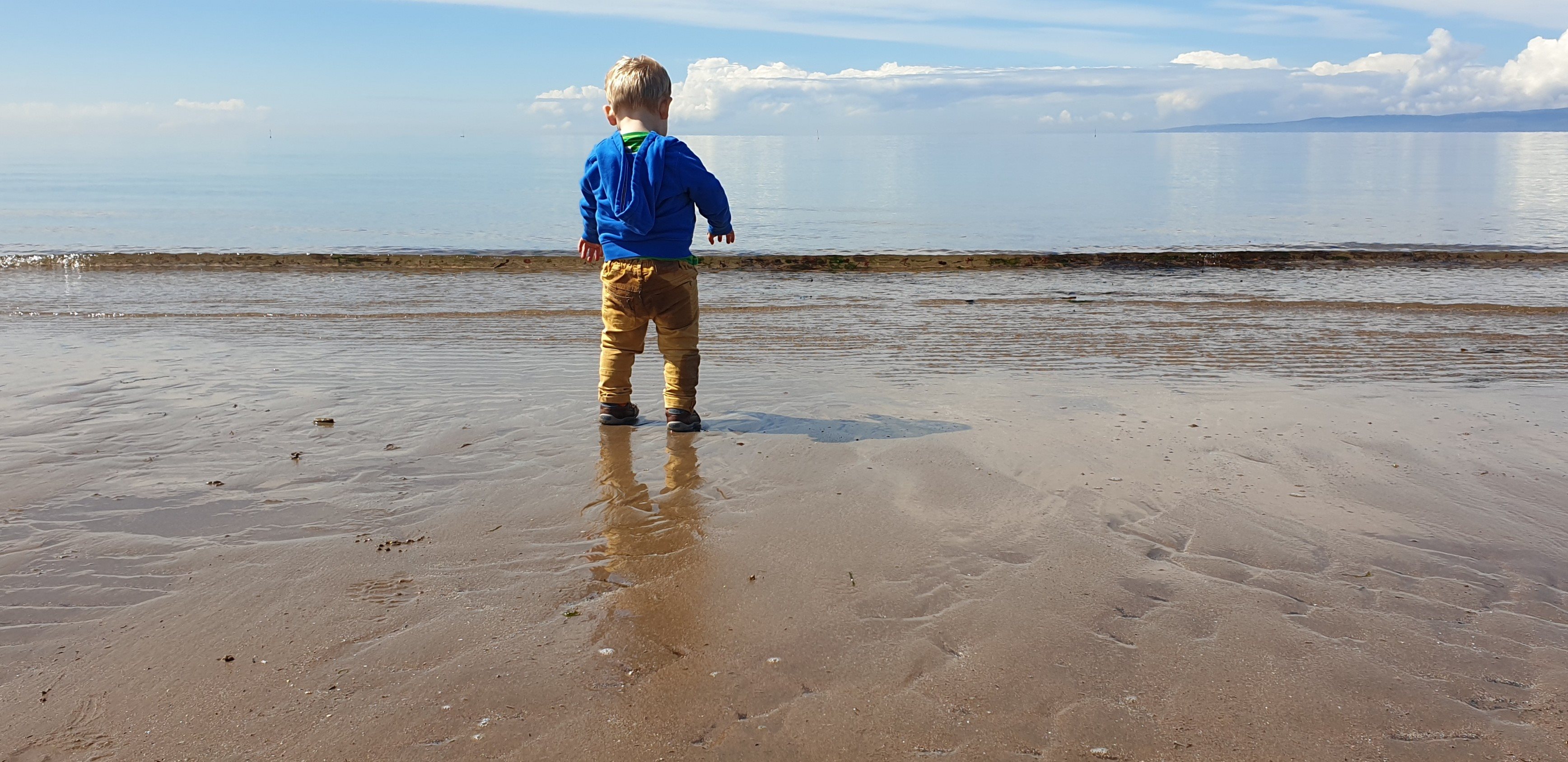 A young child with short blonde hair wearing a blue hoodie and brown pants, standing barefoot on wet sand by a calm sea under a partly cloudy sky. The child's reflection is visible on the wet sand.