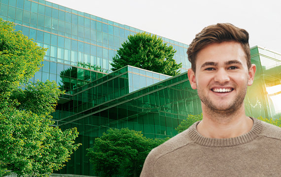Smiling young man standing in front of an aerial view of a large, green tree and building in the background.