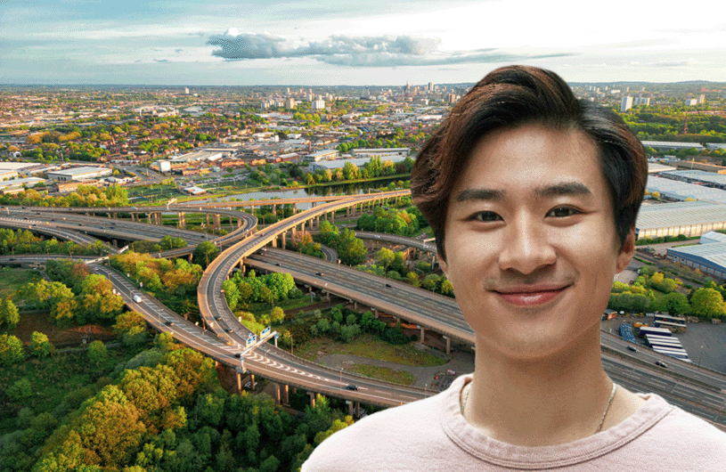 Smiling young man standing in front of an aerial view of a large, green highway interchange with a city skyline in the background.