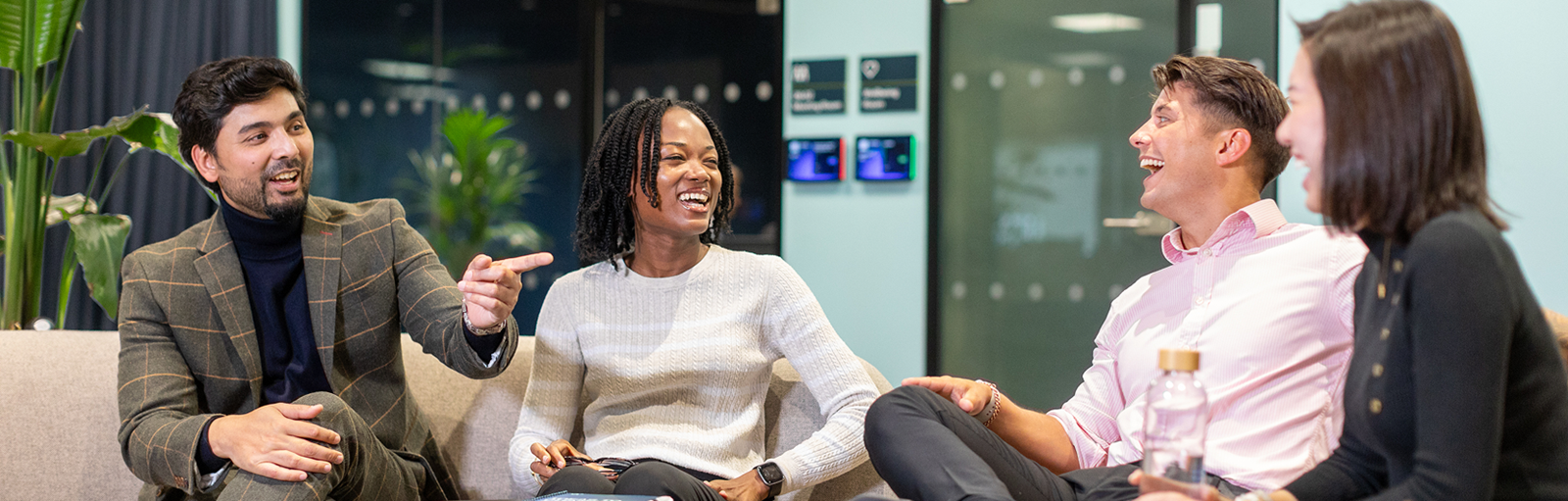 A diverse group of four young professionals sitting on a couch in a modern office lounge, laughing and having a casual conversation.