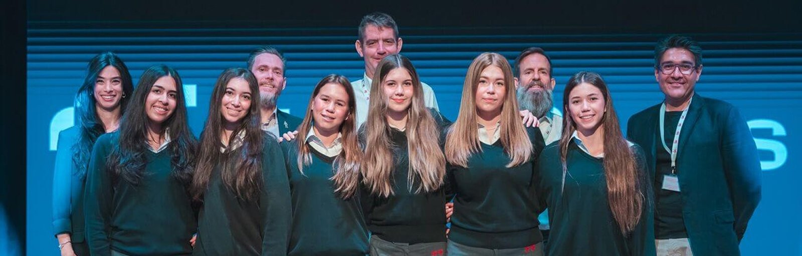 Group photo of female students and mentors on stage at an Atkins STEM event. The students are wearing matching school uniforms and are standing in front of a blue-lit backdrop. Adults stand behind them, smiling and dressed in professional attire.