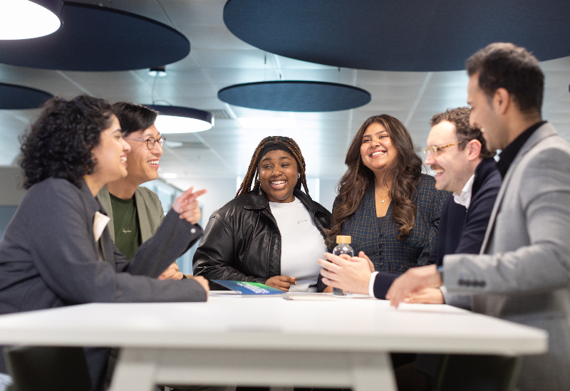 Diverse team gathered around a table in a collaborative meeting, all smiling and engaged in conversation in a contemporary workspace.