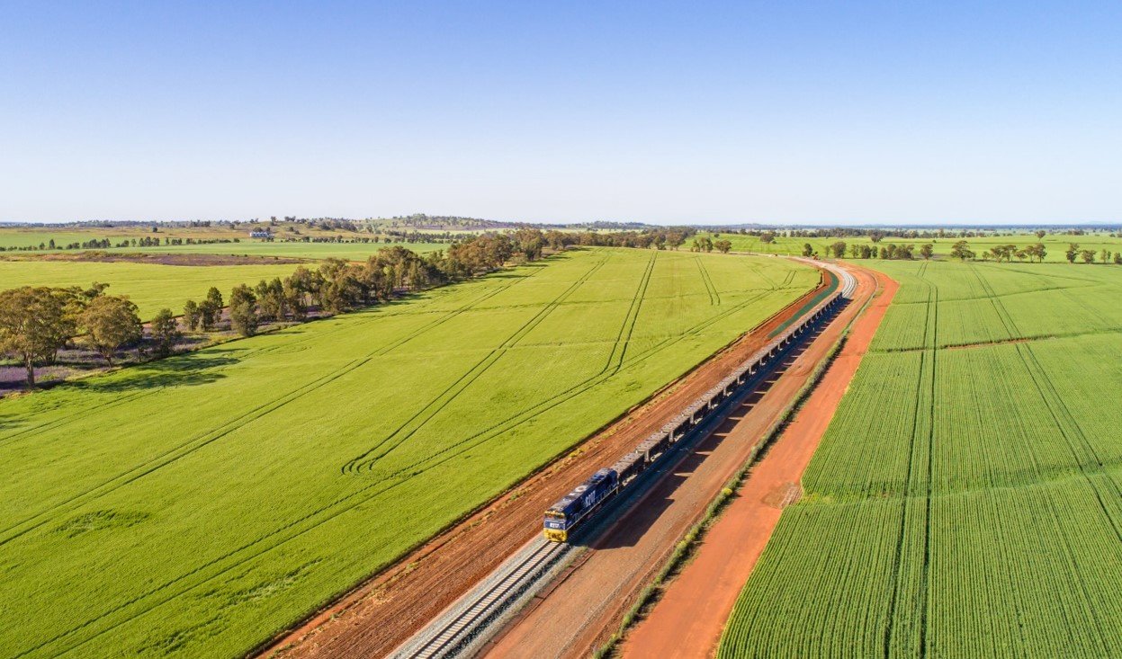 A long train moving through lush green farmland on a clear day. It appears to be an aerial (drone) shot.