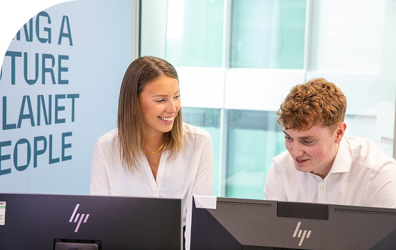 Two young professionals smiling and collaborating at their desks, with a motivational quote about the future visible on the wall behind them.