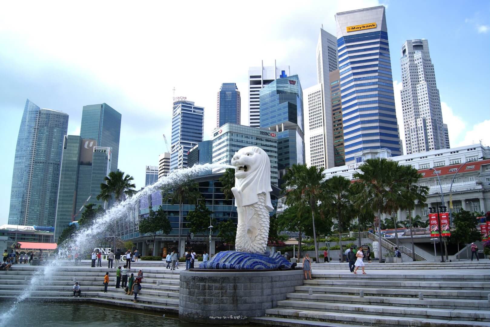 The Merlion statue in Singapore spouting water into Marina Bay, with people walking around the promenade and a backdrop of modern high-rise buildings and skyscrapers.