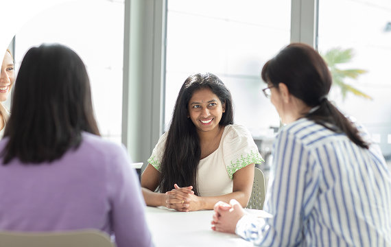 A diverse group of women smiling and having a conversation in a well-lit meeting room.