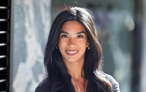A confident woman with long dark hair wearing a black shirt, standing outdoors with arms crossed and smiling, with a blurred urban background.