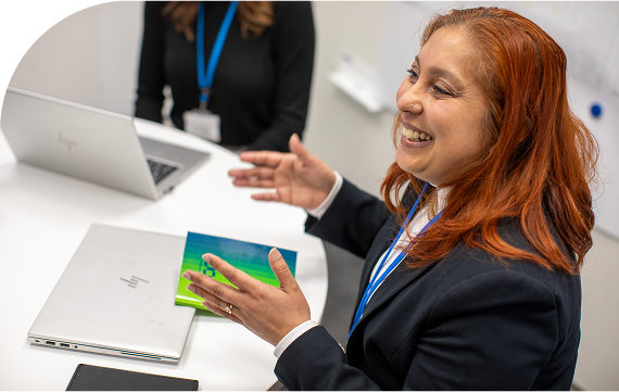 A professional woman speaking enthusiastically during a meeting, seated at a desk with a laptop.