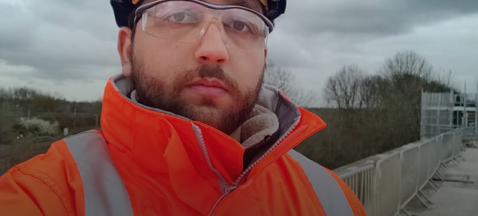 Man in high-visibility orange jacket and safety gear standing on a bridge under cloudy skies.