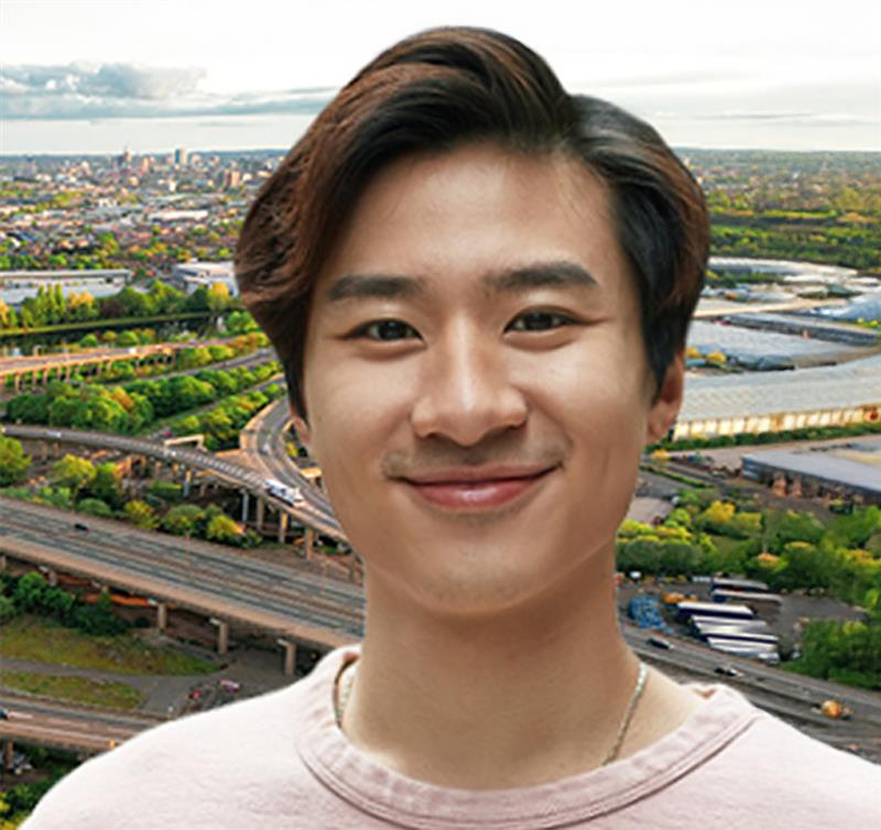 Smiling young man standing in front of an aerial view of a large, green highway interchange with a city skyline in the background