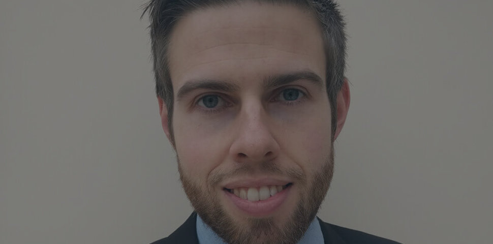 Close-up portrait of a man in a suit, smiling against a plain background.