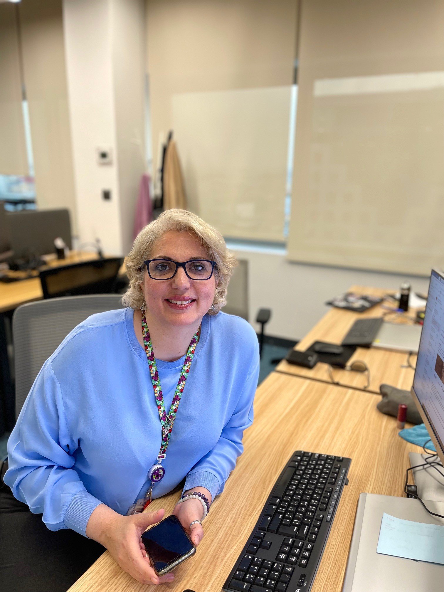 Person with short blonde hair and glasses smiling at an office desk while holding a phone.
