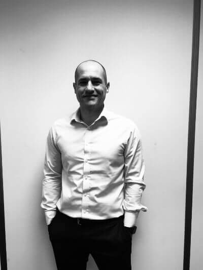 A man standing against a plain wall, wearing a light-colored dress shirt and dark trousers, with his hands in his pockets.