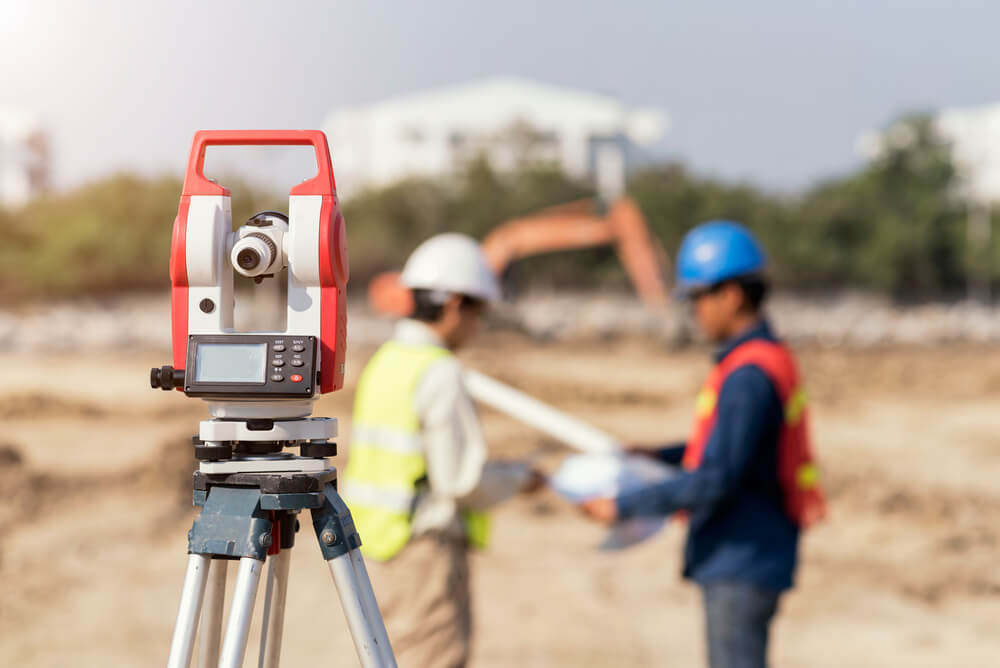 Surveying instrument on tripod at a construction site with two workers in safety gear discussing plans in the background.