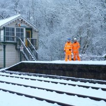 Two workers in bright orange gear standing by snowy train tracks near a small station.