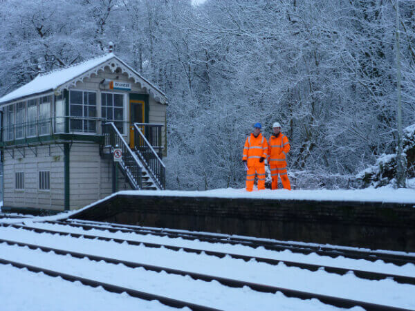 Two workers in bright orange gear standing by snowy train tracks near a small station.