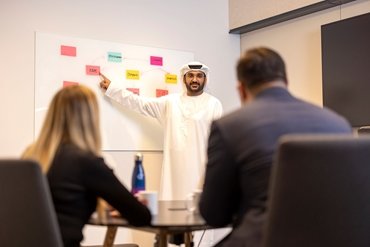 a man wearing traditional Saudi Arabian head dress and robe discussing points on a planning board with colleagues 