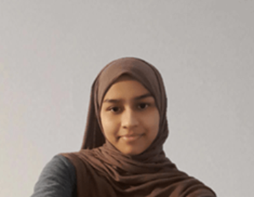 A young person wearing a brown hijab and a dark-colored top is smiling slightly while looking at the camera. The background is plain and light gray.